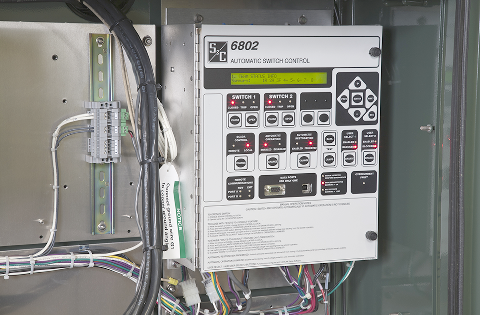 A wall-mounted 6802 Switch Control unit. The unit is on the right in the photo, while the left side shows wires affixed to the wall leading into the unit. Wires also protrude from the bottom of the unit and out of the picture frame. The 6802 Switch Control unit has an S&C logo on the upper left corner of the front panel. The front panel also has multiple labeled touchpad buttons; "Switch 1" and "Switch 2" are legible; all other text is too small to read. On some touchpad buttons, red lights are illuminated. 